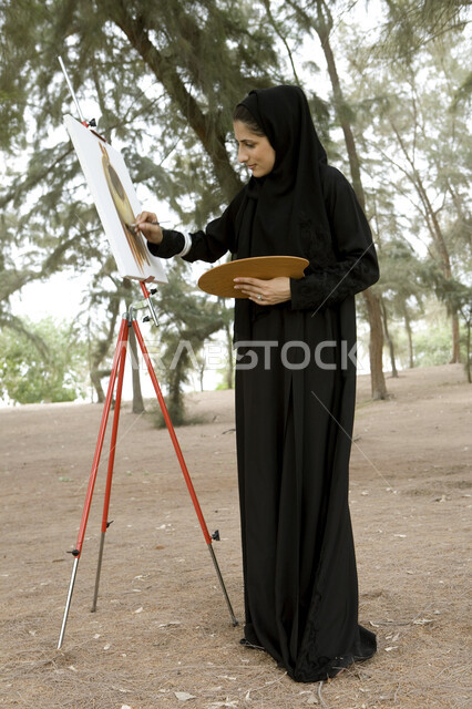 ممارسة هواية الرسم والتلوين, صورة الجسم بالطول الكامل, نشاطات ترفيهية فنية، قضاء اوقات ممتعة في إحدى الحدائق العامة, شابة عربية خليجية إماراتية محجبة تحمل بيدها بالتة ألوان وتقوم بالرسم على اللوحة في المنتزه, الإستجمام والتنزه في الهواء الطلق