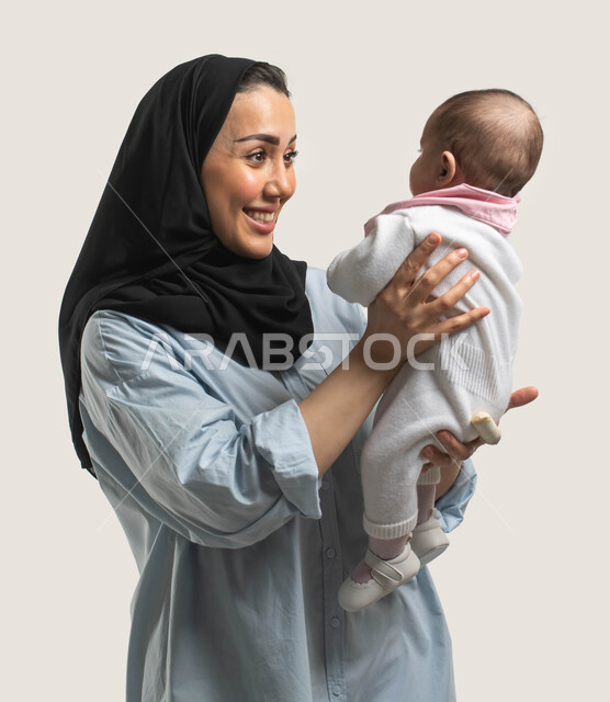 قضاء وقت ممتع مع حديثي الولاده, الاستمتاع باللعب مع الأطفال بإيماءات السرور والسعاده, بورتريه لأم عربيه سعوديه خليجيه محجبة مبتسمة ترتدي الزي الكاجوال ترفع طفلتها وتداعبها, دور الأم في حياه الأبناء, مشاعر الحب والحنان بين الأم وأولادها, خلفيه بيضاء