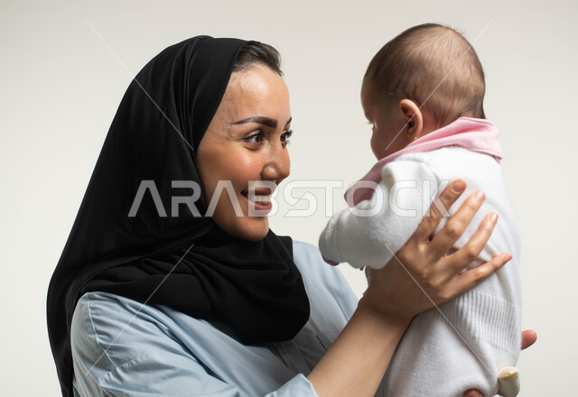 اللعب مع الأطفال بإيماءات السرور والسعاده, بورتريه مقرب لأم عربيه سعوديه خليجيه محجبة مبتسمة ترتدي الزي الكاجوال ترفع طفلتها وتداعبها, أهميه قضاء وقت ممتع مع حديثي الولاده, دور الأم في حياه الأبناء, مشاعر الحب والحنان بين الأم وأولادها, خلفيه بيضاء