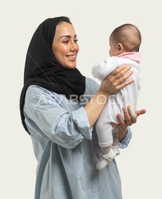 دور الأم في حياه الأبناء, الاستمتاع باللعب مع الأطفال بإيماءات السرور والسعاده, بورتريه لأم عربيه سعوديه خليجيه محجبة مبتسمة ترتدي الزي الكاجوال ترفع طفلتها وتداعبها, أهميه قضاء وقت ممتع مع حديثي الولاده, مشاعر الحب والحنان بين الأم وأولادها, خلفيه بيضاء