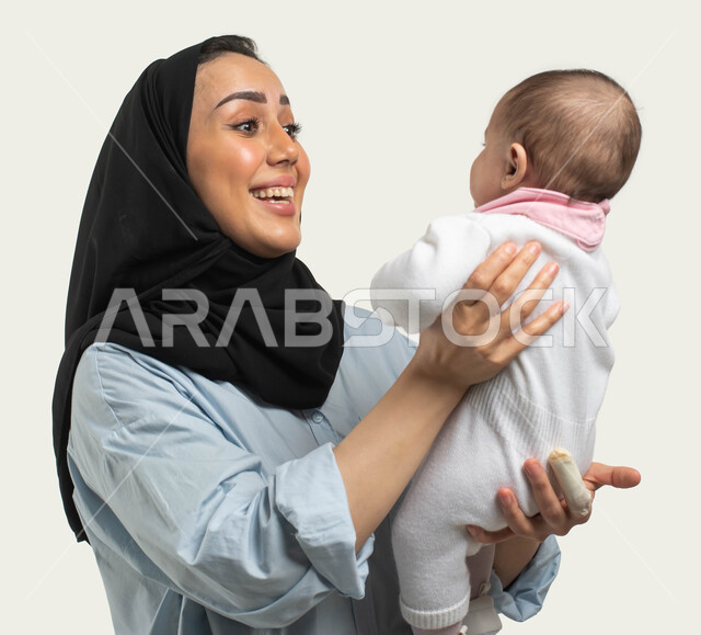 مشاعر الحب والحنان بين الأم وأولادها, الاستمتاع باللعب مع الأطفال بإيماءات السرور والسعاده, بورتريه مقرب لأم عربيه سعوديه خليجيه محجبة مبتسمة ترتدي الزي الكاجوال ترفع طفلتها وتداعبها, قضاء وقت ممتع مع حديثي الولاده, دور الأم في حياه الأبناء, خلفيه بيضاء