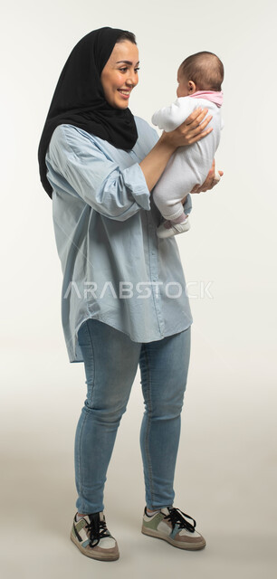 الاستمتاع باللعب مع الأطفال بإيماءات السرور والسعاده, بورتريه لأم عربيه سعوديه خليجيه محجبة مبتسمة ترتدي الزي الكاجوال ترفع طفلتها وتداعبها, أهميه قضاء وقت ممتع مع حديثي الولاد, صورة الجسم بالطول الكامل, خلفيه بيضاء