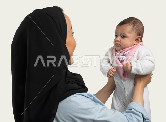 تحمل المسؤوليه ودور الأسرة السعوديه في تربيه الأبناء, قضاء أوقات ممتعة باللعب مع الأولاد بإيماءات السعادة والسرور، بورتريه مقرب لإمرأة عربية سعودية خليجية محجبة ترتدي الزي الكاجوال تحمل رضيعها بيديها لأعلى، مفهوم الأمومة والطفولة، خلفية بيضاء
