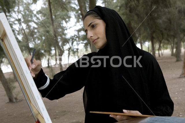 نشاطات ترفيهية فنية، قضاء اوقات ممتعة في إحدى الحدائق العامة, صورة مقربة لشابة عربية خليجية إماراتية محجبة تحمل بيدها بالتة ألوان وتقوم بالرسم على اللوحة في المنتزه, ممارسة هواية الرسم والتلوين, الإستجمام والتنزه في الهواء الطلق