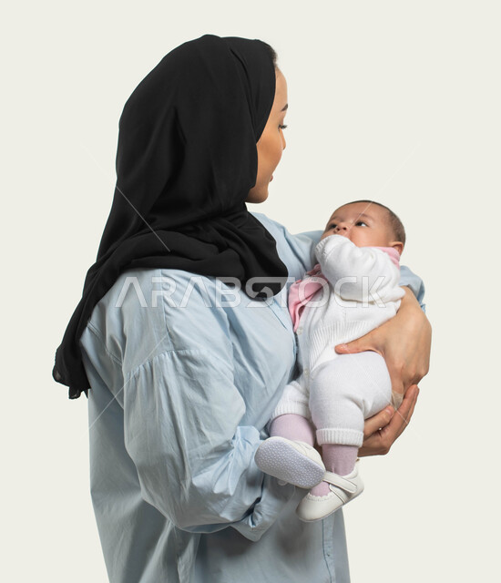 مشاعر الأم ومسؤوليتها تجاه أولادها, تقوية رابطة الأمومة بين الأم وطفلتها, بورتريه من الجانب لأم عربيه خليجيه سعوديه محجبة مبتسمة ترتدي الزي الكاجوال تحمل ابنتها بحنان وحب, النظر الى الطفله بإيماءات السرور والفرح, قضاء وقت ممتع مع الأولاد, خلفيه بيضاء