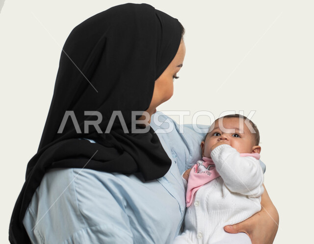 تقوية رابطة الأمومة بين الأم وطفلتها, بورتريه مقرب من الجانب لأم عربيه خليجيه سعوديه محجبة مبتسمة ترتدي الزي الكاجوال تحمل ابنتها بحنان وحب, مشاعر الأم ومسؤوليتها تجاه أولادها, النظر الى الطفله بإيماءات السرور والفرح, قضاء وقت ممتع مع الأولاد, خلفيه بيضاء