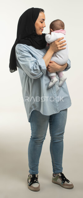 الاهتمام والعناية بالأطفال الرضع, بورتريه مقرب لأم عربيه خليجية سعوديه مبتسمة محجبة ترتدي الزي الكاجوال تقوم بحمل صغيرتها, النظر إلى الطفله بإيماءات الحب والحنان, مفهوم عاطفة الأمومه وأهميتها للأبناء, صورة الجسم بالطول الكامل, خلفيه بيضاء
