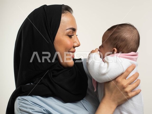 النظر إلى الطفله بإيماءات الحب والحنان, بورتريه مقرب لأم عربيه خليجية سعوديه مبتسمة محجبة ترتدي الزي الكاجوال تقوم بملاطفه ومداعبه ابنتها, مفهوم عاطفة الأمومه وأهميتها للأبناء, دور الأسرة السعوديه في الاهتمام والعناية بالأطفال الرضع, خلفيه بيضاء