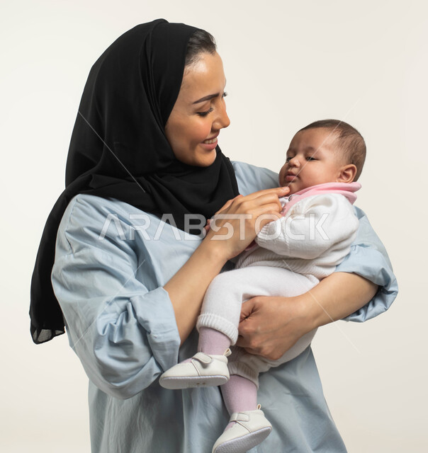 الرعاية والاهتمام بالأبناء, النظر إلى الطفلة بإيماءات الحب والحنان, بورتريه لأم عربيه خليجيه سعوديه مبتسمة محجبة ترتدي الزي الكاجوال تقوم بإضحاك ابنتها وملاعبتها, طفلة رضيعه ترتدي بربتوز أبيض مريح بملامح طفوليه بريئة, خلفيه بيضاء
