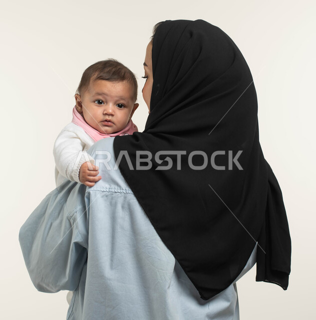 مشاعر العطف والحنان, بورتريه مقرب من الخلف لأم عربيه خليجيه سعوديه ترتدي الزي الكاجوال تحمل طفلتها بحب ولطف, طفلة رضيعه تنظر إلى شيء ما بإيماءات الطفولة والبراءة, مساعده الأبناء والبنات على الاسترخاء والهدوء, الاهتمام بالأطفال ورعايتهم, خلفيه بيضاء
