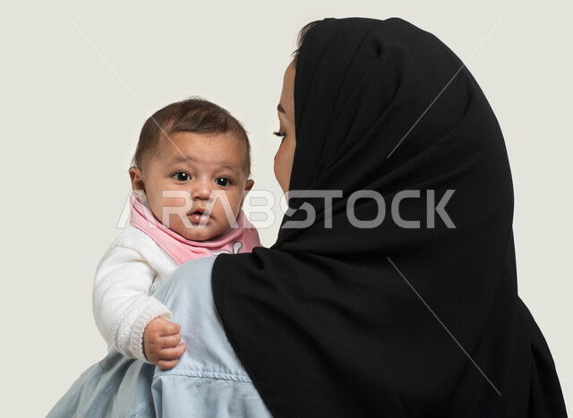 اهتمام الأمهات بالأطفال ورعايتهم, بورتريه مقرب من الخلف لإمرأة عربيه خليجيه سعوديه ترتدي الزي الكاجوال تحمل طفلتها بحب وحنان, طفلة رضيعه تنظر إلى شيء ما بإيماءات الطفولة والبراءة, مساعده الأبناء والبنات على الاسترخاء والهدوء, خلفيه بيضاء