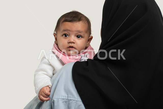 الاهتمام بالأطفال ورعايتهم, بورتريه مقرب من الخلف لإمرأة عربيه خليجيه سعوديه ترتدي الزي الكاجوال تحمل طفلتها بحب وحنان, طفلة رضيعه تنظر إلى شيء ما بإيماءات الطفولة والبراءة, مساعده الأبناء والبنات على الاسترخاء والهدوء, خلفيه بيضاء