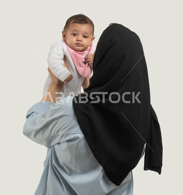 أهميه قضاء وقت ممتع مع حديثي الولاده, الاستمتاع باللعب مع الأطفال, بورتريه من الخلف لأم عربيه سعوديه خليجيه محجبة ترتدي الزي الكاجوال ترفع طفلها للأعلى, دور الأم في حياه الأبناء, مشاعر الحب والحنان بين الأم وأولادها, خلفيه بيضاء
