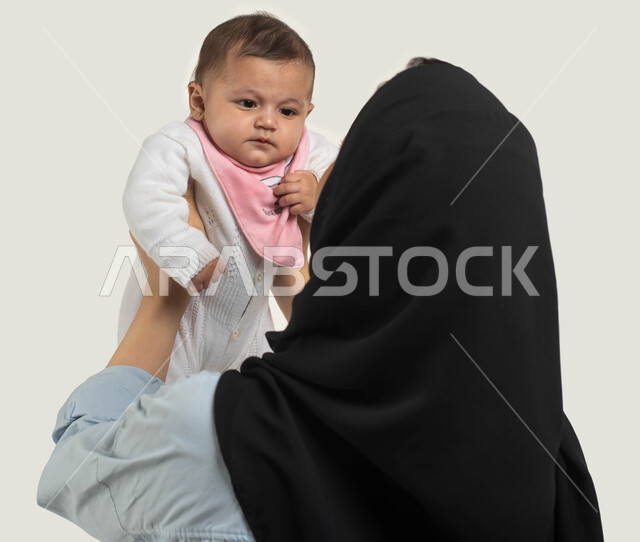 الاستمتاع باللعب مع الأطفال, بورتريه مقرب من الخلف لأم عربيه سعوديه خليجيه محجبة ترتدي الزي الكاجوال ترفع طفلها للأعلى, أهميه قضاء وقت ممتع مع حديثي الولاده, دور الأم في حياه الأبناء, مشاعر الحب والحنان بين الأم وأولادها, خلفيه بيضاء