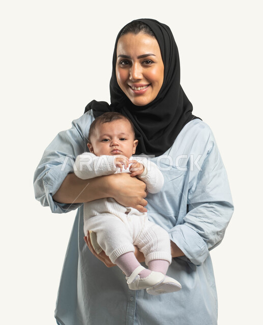 الوقوف والنظر إلى الكاميرا بإيماءات السعادة والسرور, بورتريه لأم عربيه خليجية سعودية محجبة مبتسمة ترتدي الزي الكاجوال تحمل طفلتها الرضيعه, مفهوم عاطفه الأمومه وأهميتها للطفل, تقوية الرابط الأسري بين الأهل والأبناء, خلفيه بيضاء