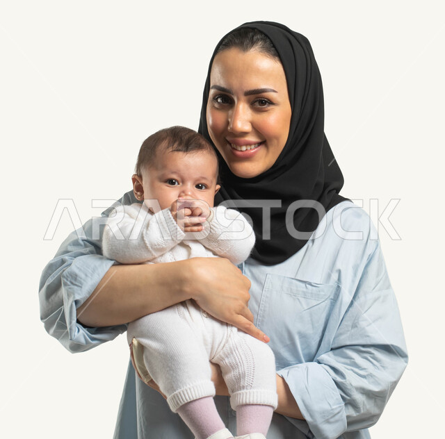 مفهوم عاطفه الأمومه وأهميتها للطفل, الوقوف والنظر إلى الكاميرا بإيماءات السعادة والسرور, بورتريه لأم عربيه خليجية سعودية محجبة مبتسمة ترتدي الزي الكاجوال تحمل طفلتها الرضيعه, تقوية الرابط الأسري بين الأهل والأبناء, خلفيه بيضاء