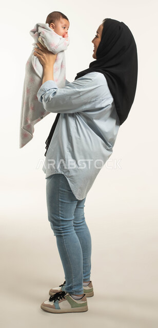 Enjoying spending quality time with children, looking at the newborn with gestures of tenderness and love, side portrait of a Saudi Arabian Gulf mother wearing a casual outfit, caressing her child, the role of parents in developing children’s sensory and motor skills, full-length body image, white background