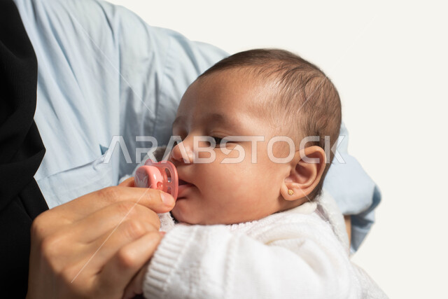 مشاعر الحنان والعطف, دور الأم في العناية بأبنائها, صورة مقربة لامرأة عربية خليجية سعوديه تحمل رضيعتها بحب وتقوم بوضع اللهاية لها, استخدام الادوات لاسترخاء وتهدئة الأطفال الرضع, مفهوم الطفولة والأمومة, خلفيه بيضاء