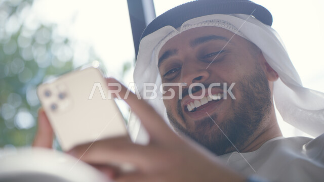 التسويق الالكتروني الذكي والآمن، دردشات ومحادثات عبر مواقع التواصل الاجتماعي, صورة مقربة لرجل عربي خليجي إماراتي مبتسم يرتدي الكندورة والغترة يستخدم الهاتف المحمول, التواصل مع الأصدقاء عبر الانترنت بإيماءات الفرح والسرور
