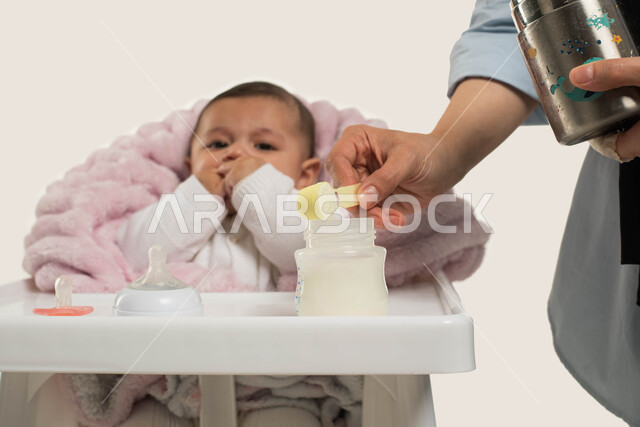 الجلوس في كرسي الطعام المخصص, بورتريه مقرب لطفلة عربيه خليجية سعوديه تضع يدها في فمها بإيماءات الجوع, أم تقوم بتحضير وتجهيز الحليب الصناعي لطفلتها, اهتمام الأم بصغارها والعنايه بهم, مفهوم الأمومه والطفوله, خلفيه بيضاء