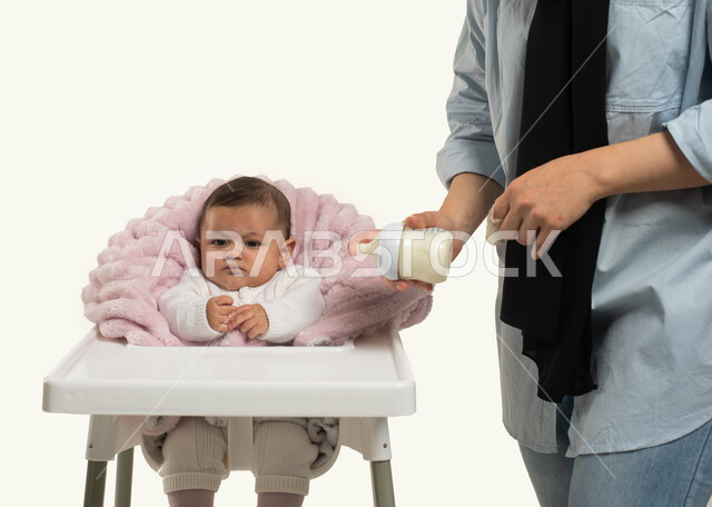 مسؤوليه الأم تجاه أولادها, بورتريه مقرب لطفله رضيعه عربيه خليجية سعوديه تجلس في كرسي الطعام المخصص, أم ترتدي الزي الكاجوال تقوم بتحضير الحليب لطفلتها, مفهوم الرضاعه الصناعيه, الاهتمام بتغذية ونمو الأطفال, خلفيه بيضاء