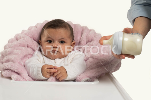 دور ومسؤوليه الأم تجاه أولادها, بورتريه مقرب لطفله رضيعه عربيه خليجية سعوديه تجلس في كرسي الطعام المخصص, أم ترتدي الزي الكاجوال تقوم بتحضير الحليب لطفلتها, مفهوم الرضاعه الصناعيه, الاهتمام بتغذية ونمو الأطفال, خلفيه بيضاء