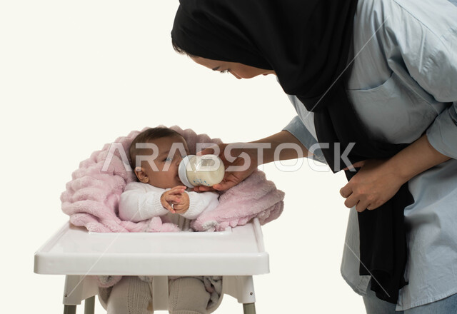 مفهوم الأمومه والرضاعه الصناعيه, الاهتمام بتغذية ونمو الأطفال, بورتريه من الجانب لأم عربيه خليجيه سعوديه ترتدي الزي الكاجوال تقوم بإعطاء طفلتها الحليب الصناعي من عبوه الرضاعه, طفلة رضيعة تجلس في كرسي الطعام, دور وحنان الأم في رعايه أبنائها, خلفيه بيضاء