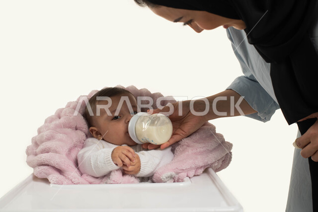 الوقوف والنظر إلى الطفله بإيماءات العطف والحنان, صورة مقربه لأم عربية خليجية سعوديه محجبة تقوم بإعطاء طفلتها الحليب الصناعي من عبوه الرضاعه, طفله رضيعه تجلس في كرسي الطعام المخصص, دور وحنان الأم في رعايه أبنائها, خلفيه بيضاء