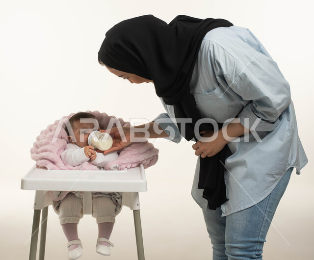 الاهتمام بتغذية ونمو الأطفال, بورتريه من الجانب لأم عربيه خليجيه سعوديه ترتدي الزي الكاجوال تقوم بإعطاء طفلتها الحليب الصناعي من عبوه الرضاعه, طفلة رضيعة تجلس في كرسي الطعام, مفهوم الأمومه والرضاعه الصناعيه, دور وحنان الأم في رعايه أبنائها, خلفيه بيضاء