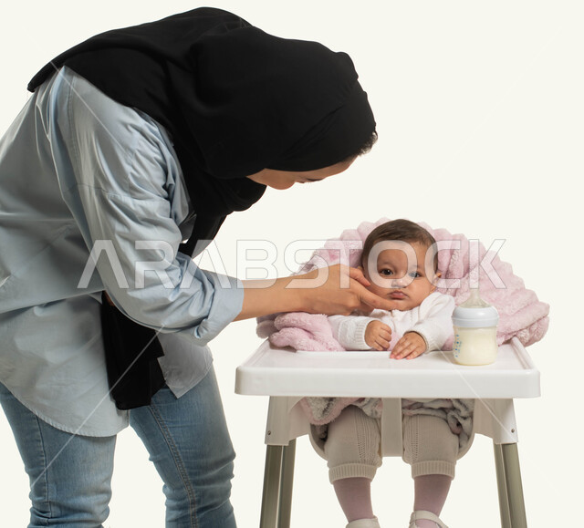 ايماءات اللعب والحب والسعادة ,الاعتناء والاهتمام بتغذية الأبناء, بورتريه مقرب لأم عربية خليجيه سعودية محجبة ترتدي الزي الكاجوال تقف بجانب طفلتها الصغيرة، جلوس الرضيع على كرسي الطعام، تناول الحليب من عبوة الرضاعه, قضاء أوقات ممتعه مع الأطفال, خلفية بيضاء