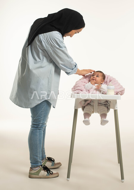 Gently caressing with feelings of love and tenderness, a close-up portrait of a veiled Saudi Arabian Gulf mother wearing a casual outfit standing next to her little girl, the infant sitting on the dining chair, drinking milk from a feeding bottle, spending enjoyable times with the children, full-length body portrait, white background