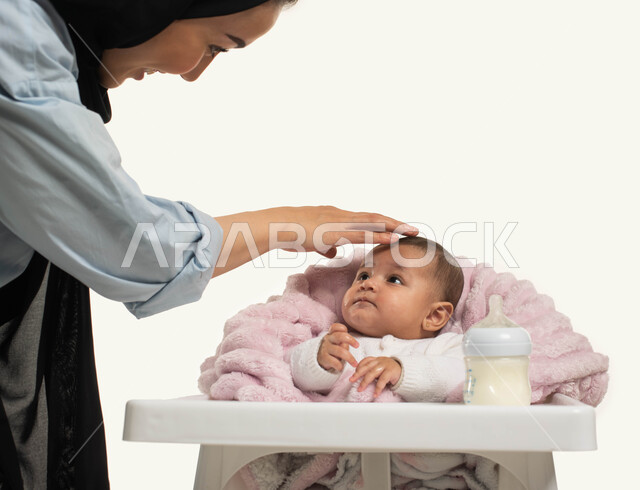 تناول الحليب من عبوة الرضاعه, المداعبه بلطف مع مشاعر الحب والحنان, بورتريه مقرب لأم عربية خليجيه سعودية محجبة ترتدي الزي الكاجوال تقف بجانب طفلتها الصغيرة، جلوس الرضيع على كرسي الطعام، قضاء أوقات ممتعه مع الأطفال, خلفية بيضاء