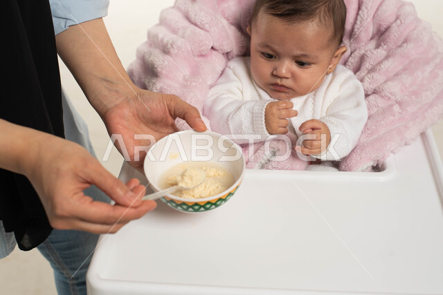 Family and responsibility, the concept of caring and taking care of children’s nutrition, strengthening the maternal bond between the mother and her child, a close-up portrait of a Saudi Gulf Arab baby girl sitting on a dining chair, a Saudi Gulf Arab woman feeding her daughter a meal, white background
