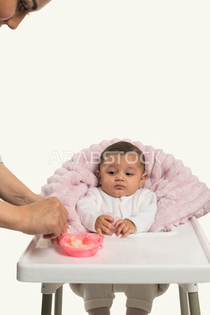 مفهوم الاهتمام و الرعياه بتغذية الأبناء, تقوية رابطة الامومة بين الام وطفلتها, بورتريه مقرب لطفلة رضيعه عربيه خليجية سعودية تجلس على كرسي الطعام، سيده عربيه خليجيه سعودية تقوم بإطعام ابنتها وجبه الطعام, قضاء أوقات ممتعه مع الأطفال, خلفية بيضاء