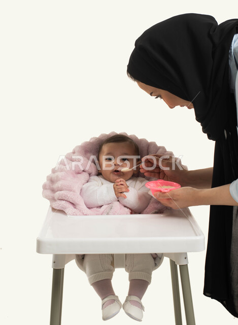 تقوية رابطة الامومة بين الام وطفلتها, بورتريه مقرب لسيده عربيه خليجيه سعودية محجبه تقوم بإطعام ابنتها وجبه الطعام, جلوس الرضيع على كرسي الطعام، مفهوم الرعايه والاهتمام بتغذية الأبناء, قضاء أوقات ممتعه مع الأطفال, خلفية بيضاء