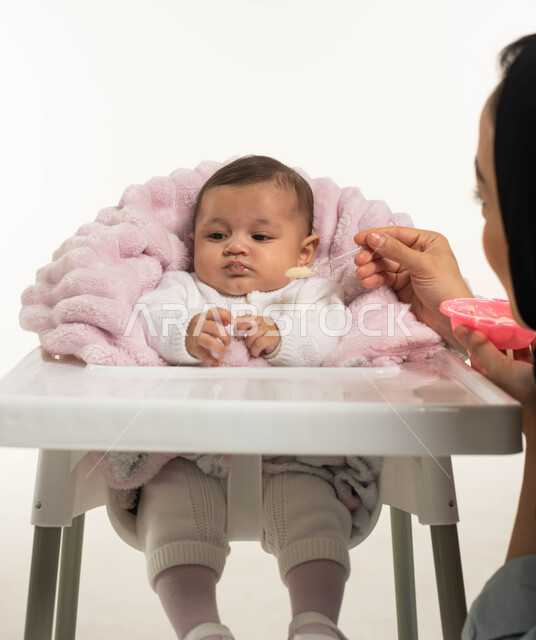 مفهوم الرعايه والاهتمام بتغذية الأبناء, تقوية رابطة الامومة بين الام وطفلتها, بورتريه مقرب لسيده عربيه خليجيه سعودية محجبه تقوم بإطعام ابنتها وجبه الطعام, جلوس الرضيع على كرسي الطعام، قضاء أوقات ممتعه مع الأطفال, خلفية بيضاء