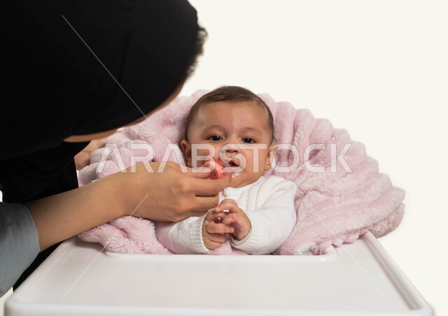 حنان الأم وعطفها على أطفالها, الرعايه والاهتمام بالأبناء, بورتريه مقرب لسيده عربية خليجيه سعودية محجبة ترتدي الزي الكاجوال تقوم بإعطاء طفلتها الصغيرة اللهاية، جلوس الرضيع على كرسي الطعام, قضاء أوقات ممتعه مع الأطفال, خلفية بيضاء