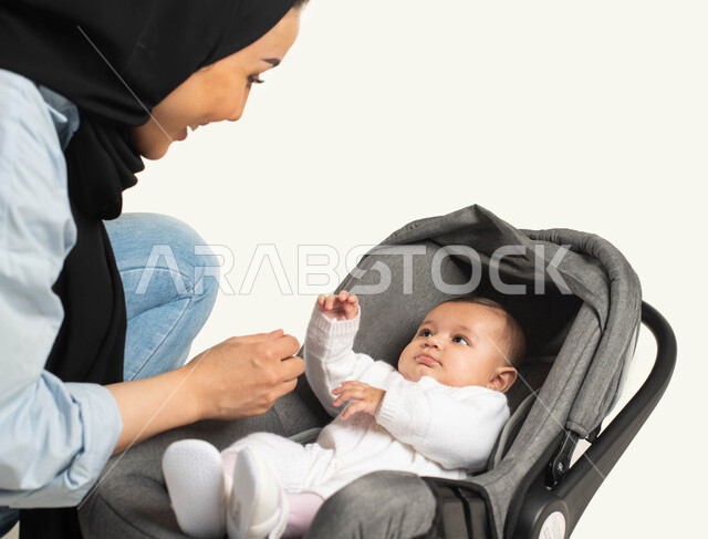 اللعب مع الأبناء بإيماءات الحب والأمان، الاهتمام والاعتناء بالأطفال ، بورتريه مقرب لام عربية خليجيه سعودية محجبة ترتدي الزي الكاجوال تجلس بجانب طفلها الصغير، جلوس الرضيع في الكرسي المخصص له, قضاء أوقات ممتعه مع الأطفال, خلفية بيضاء