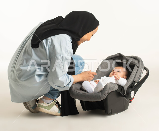 جلوس الرضيع في الكرسي المخصص له, قضاء أوقات ممتعه مع الأطفال, اللعب مع الأبناء بإيماءات الحب والأمان، الاهتمام والاعتناء بالأطفال ، بورتريه لام عربية خليجيه سعودية محجبة ترتدي الزي الكاجوال تجلس بجانب طفلها الصغير، خلفية بيضاء