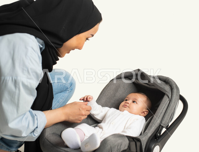  قضاء أوقات ممتعه مع الأطفال, اللعب مع الأبناء بإيماءات الحب والأمان، الاهتمام والاعتناء بالأطفال ، بورتريه مقرب لام عربية خليجيه سعودية محجبة ترتدي الزي الكاجوال تجلس بجانب طفلها الصغير، جلوس الرضيع في الكرسي المخصص له, خلفية بيضاء