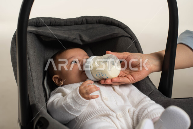 Feelings of tenderness and kindness, concern and care for children’s nutrition, a Saudi mother giving her child useful food, a close-up portrait of a Saudi Arabian Gulf girl sitting in her own chair and drinking milk from a breastfeeding bottle, strengthening the maternal bond, white background.