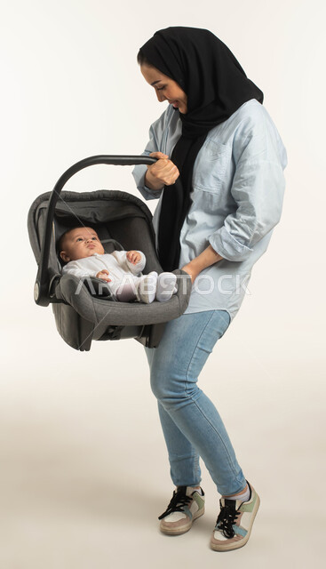 النظر إلى الابنه بإيماءات الحنان والحب, دور الأم وأهميتها الأساسي في تربية ونشأة الأطفال، بورتريه لسيدة عربية خليجيه سعودية مبتسمة محجبة ترتدي الزي الكاجوال تحمل كرسي طفلتها الرضيعه , صورة الجسم بالطول الكامل, خلفية بيضاء