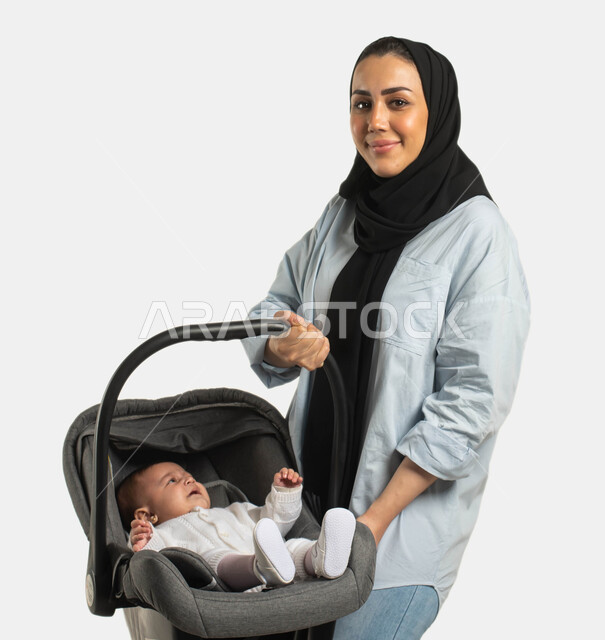 أهمية الأم ودورها الأساسي في تربية ونشأة الأطفال, مفهوم الأمومة والرعايه والاهتمام بالأبناء ، بورتريه لسيدة عربية خليجيه سعودية مبتسمة محجبة ترتدي الزي الكاجوال تحمل كرسي طفلتها الرضيعه و تنظر إلى الكاميرا بإيماءات السعادة والحب, خلفية بيضاء