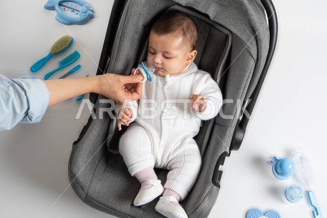  تقوية رابطة الأمومة , الاهتمام والرعاية بالأبناء, أم سعودية تقوم بإعطاء طفلها اللهاية ، بورتريه لطفلة عربية خليجية سعودية لطيفة ترتدي لباس أبيض قطني تجلس في الكرسي الخاص بها ،مشاعر العطف والحنان , خلفية بيضاء