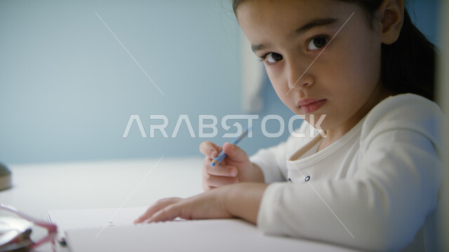 الدراسة في الإمارات العربية المتحدة، صورة مقربة لفتاة صغيرة عربية خليجية إماراتية تجلس على الطاولة وتقوم بأداء الواجبات المدرسية، التعليم المنزلي أثناء الحجر الصحي و تفشي فايروس كورونا ، التعليم عن بعد, النظر إلى الكاميرا