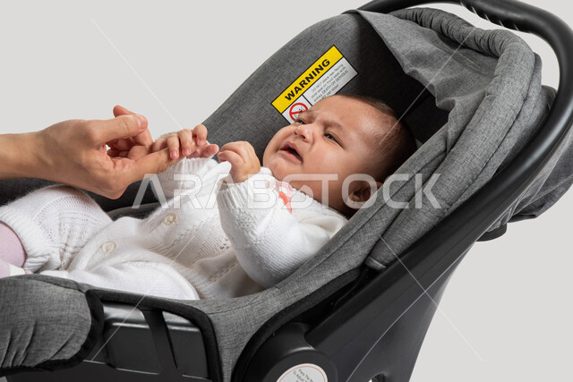 الاعتناء والاهتمام بالأبناء و رعايتهم, ايماءات البكاء و الشعور بالانزعاج ,أم سعودية تحاول مساعدة طفلتها على تنمية الحواس , بورتريه مقرب لطفلة عربيه خليجيه سعودية لطيفة ترتدي لباس أبيض قطني تجلس في كرسيها الخاص , خلفية بيضاء