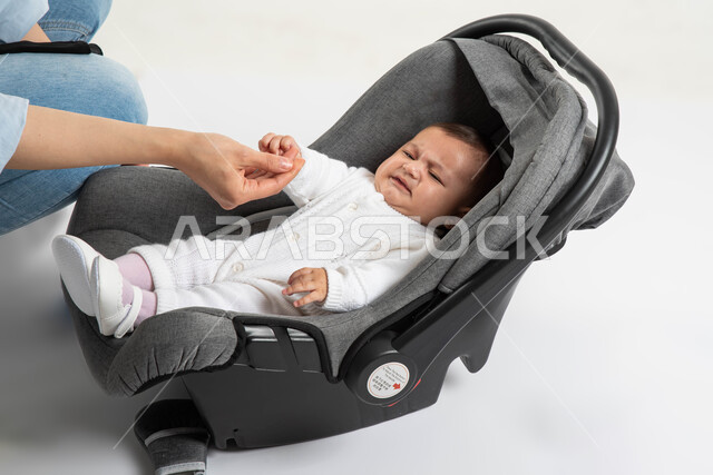 ايماءات البكاء و الشعور بالانزعاج ,أم سعودية تحاول مساعدة طفلتها على تنمية الحواس , الاهتمام والاعتناء بالأبناء و رعايتهم, بورتريه لطفلة عربيه خليجيه سعودية لطيفة ترتدي لباس أبيض قطني تجلس في كرسيها الخاص , خلفية بيضاء