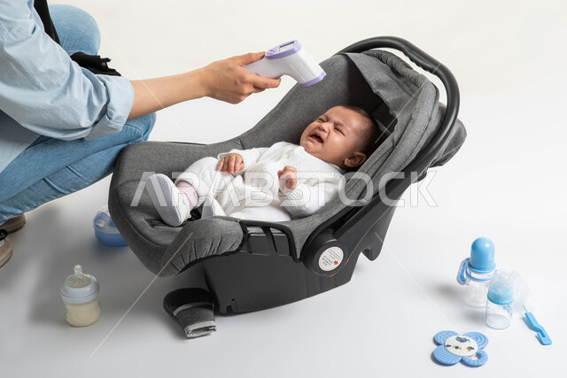 الاهتمام والاعتناء بالأبناء و رعايتهم, ايماءات البكاء و الشعور بالتعب و المرض, أدوات الرضاعه ومستلزمات الرضيع, أم سعودية تقوم بفحص حرارة طفلتها بالجهاز الرقمي, بورتريه لطفلة عربيه خليجيه سعودية لطيفة ترتدي بربتوز أبيض قطني تجلس في كرسيها الخاص, خلفية بيضاء