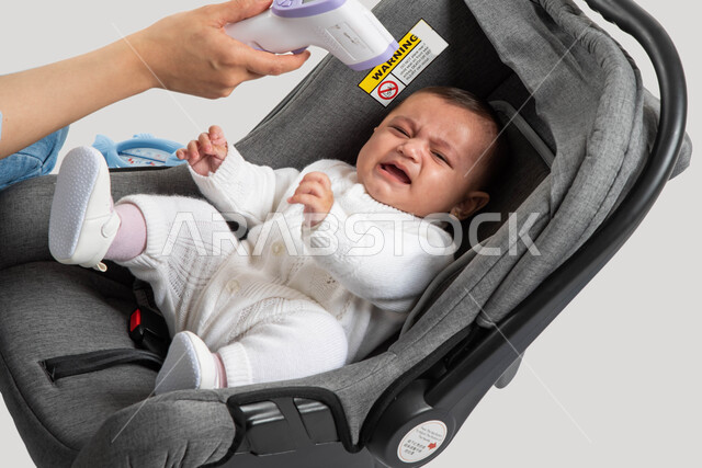 ايماءات البكاء و الشعور بالتعب و المرض, أم سعودية تقوم بفحص حرارة طفلتها بالجهاز الرقمي, الاهتمام والاعتناء بالأبناء و رعايتهم, بورتريه لطفلة عربيه خليجيه سعودية لطيفة ترتدي بربتوز أبيض قطني تجلس في كرسيها الخاص, خلفية بيضاء