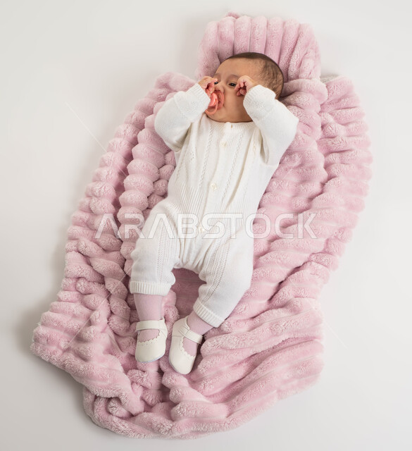 استخدام اللهاية للاسترخاء والنوم, بورتريه لطفلة عربيه خليجيه سعودية لطيفة ترتدي بربتوز أبيض قطني مستلقية على غطاء فرو باللون الوردي, الاهتمام بصحة الأبناء والعناية بهم, خلفية بيضاء